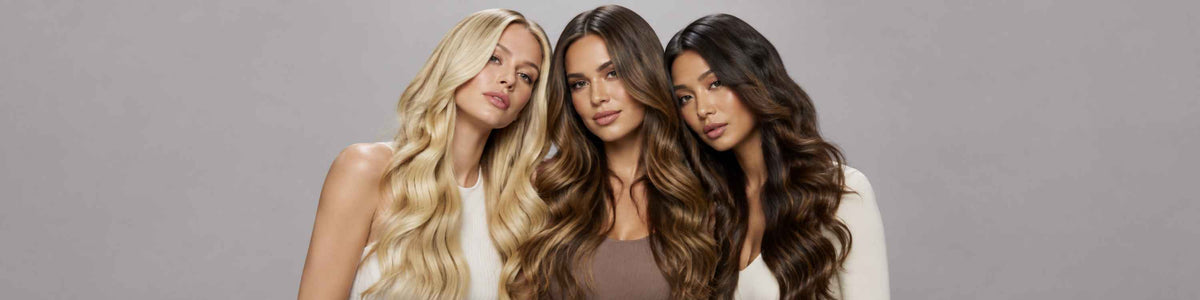 Three women with long, wavy hair in blonde, brown, and dark brown tones, posing together against a solid gray studio background.